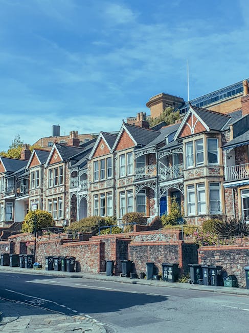 A row of Victorian-style terraced houses on a residential street, featuring ornate decorative ironwork on balconies and verandas, with large bay windows framed in white paint. The buildings are constructed from red brick with contrasting light-colored stone or painted details, and each house has a small front garden separated from the pavement by low brick walls, some with shrubbery or small trees. The street is lined with black wheelie bins placed along the curb, indicative of privately managed waste collection, possibly for recycling and general rubbish. Clear blue sky above suggests a bright day, with the sunlight casting soft shadows on the buildings and highlighting their architectural details. In the background, modern structures are visible, blending Victorian and contemporary urban environments, which reflects the local context of private waste handling and the use of external rubbish removal services such as Waste Removal Bromley for larger or special waste loads. The scene emphasizes a typical residential setting where or alternative rubbish disposal methods are employed to maintain cleanliness and manage waste effectively.