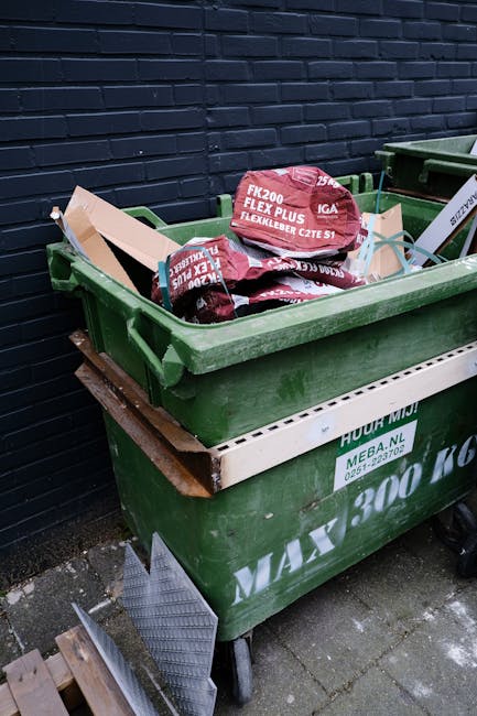 A green wheeled waste bin filled with red and black bags of garden or landscaping materials, including flexible garden hoses, placed on a paved outdoor area adjacent to a dark brick wall. The bin is situated on top of a wooden pallet, and there is a second similar bin partially visible behind it. A white plastic barrier or trim is attached to the front of the bin, with some printed text, and a small metal ramp or grate rests against the lower side of the bin. The scene suggests on-site waste collection or an alternative rubbish removal method, with Waste Removal Bromley providing local rubbish disposal services in a residential or commercial setting. Natural daylight illuminates the scene, highlighting the textures of the plastic, bricks, and paving stones, with no visible recycling or disposal equipment besides the wheeled bins, indicating typical private waste handling outside of municipal collection.