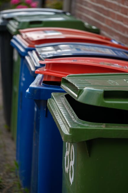 A row of large, rectangular wheelie bins lined up outdoors along a paved pathway adjacent to a brick wall. The bins are made of durable plastic with textured surfaces, featuring prominent lids that are secured closed. The bins are in various colours: green, blue, red, and dark green, each with a matte or slightly glossy finish. The green bin in the foreground has a white recycling symbol on its side. The lids are neatly aligned, with some slightly open or resting on the top edges, and the bins are positioned tightly together, extending into the background. The outdoor setting is indicated by the presence of natural light, and the scene appears to depict an area designated for waste collection or recycling, consistent with private waste handling or rubbish removal services such as those offered by Waste Removal Bromley. The environment suggests a residential or commercial location with a clean, orderly appearance, highlighting the typical use of these bins for segregated waste disposal and alternative rubbish collection methods.