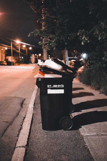 A green wheeled waste bin filled with red and black bags of garden or landscaping materials, including flexible garden hoses, placed on a paved outdoor area adjacent to a dark brick wall. The bin is situated on top of a wooden pallet, and there is a second similar bin partially visible behind it. A white plastic barrier or trim is attached to the front of the bin, with some printed text, and a small metal ramp or grate rests against the lower side of the bin. The scene suggests on-site waste collection or an alternative rubbish removal method, with Waste Removal Bromley providing local rubbish disposal services in a residential or commercial setting. Natural daylight illuminates the scene, highlighting the textures of the plastic, bricks, and paving stones, with no visible recycling or disposal equipment besides the wheeled bins, indicating typical private waste handling outside of municipal collection.