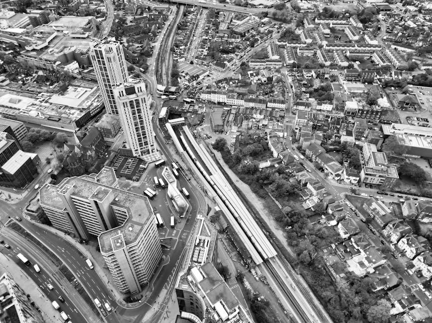A black-and-white aerial photograph of a dense urban area showing a variety of buildings, including high-rise office towers with flat roofs and glass facades, low-rise residential houses with pitched roofs, and commercial structures. In the foreground, there are multiple multi-story office blocks arranged around a road junction with parked cars and vans nearby. A railway line runs diagonally through the middle of the image, with railway tracks visible on elevated and ground-level sections, and some train cars parked along the tracks. Surrounding the railway, there are patches of greenery and trees interspersed among the buildings. The background reveals a network of roads, bridges, and additional urban development, with more rooftops, streets, and clusters of smaller residential buildings extending into the distance. This scene emphasizes the dense layout of a city with varied architectural styles and a significant transportation infrastructure, which waste removal services like those provided by Waste Removal Bromley support by managing waste generated throughout the area, including commercial, residential, and transportation-related refuse.