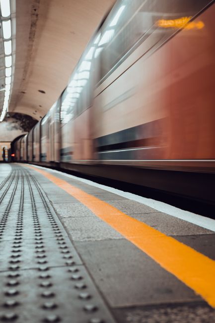 The image depicts a scene inside a modern underground train station with a train approaching the platform. The train has a sleek, streamlined design with a silver and orange-brown exterior, reflecting the station's lighting. The side of the train features horizontal black and silver stripes, and the windows are tinted, appearing as dark longitudinal bands. The platform is made of smooth, grey concrete with textured tactile paving along the edge, featuring small, raised bumps for safety, and painted with a bright orange safety line running parallel to the train. The background shows the curved ceiling of the tunnel, illuminated by overhead lighting that creates a soft, warm glow, while the tunnel’s sides are lined with light-colored, smooth panels. The overall environment appears clean and maintained, supporting the context of efficient public transit or alternative transportation infrastructure, with the mention of waste removal referencing the importance of keeping such spaces tidy and free of debris, as managed by companies like Waste Removal Bromley.